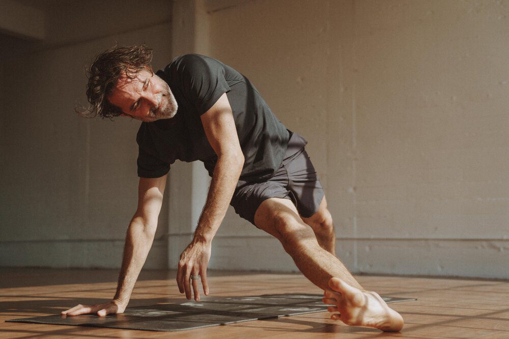 Yoga instructor and The Light Orchard founder Dalton Grant in a black athletic outfit does a yoga flow on a yoga mat in a sunlit room.